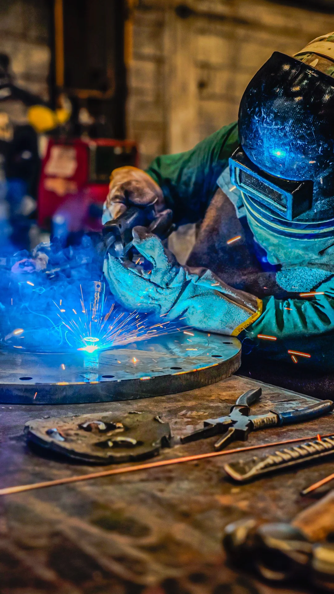 Welder in a full welding helmet and gloves welding a round metal plate on a workbench, with sparks and tools around it