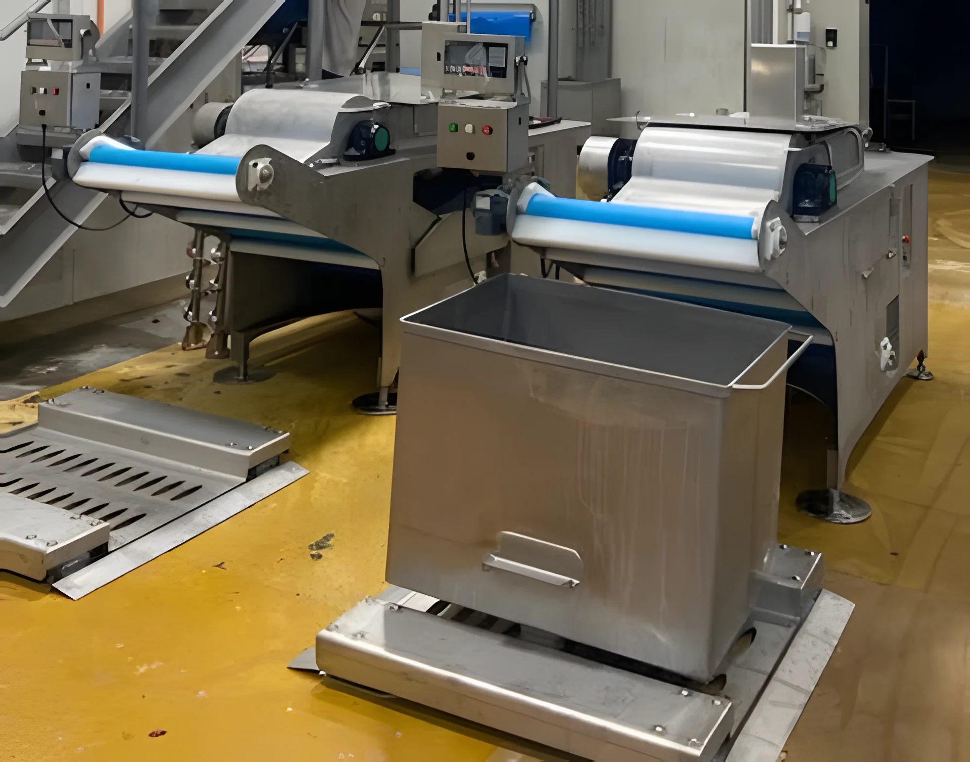 Two stainless steel processing machines with conveyor belts and a stainless bin on a factory floor