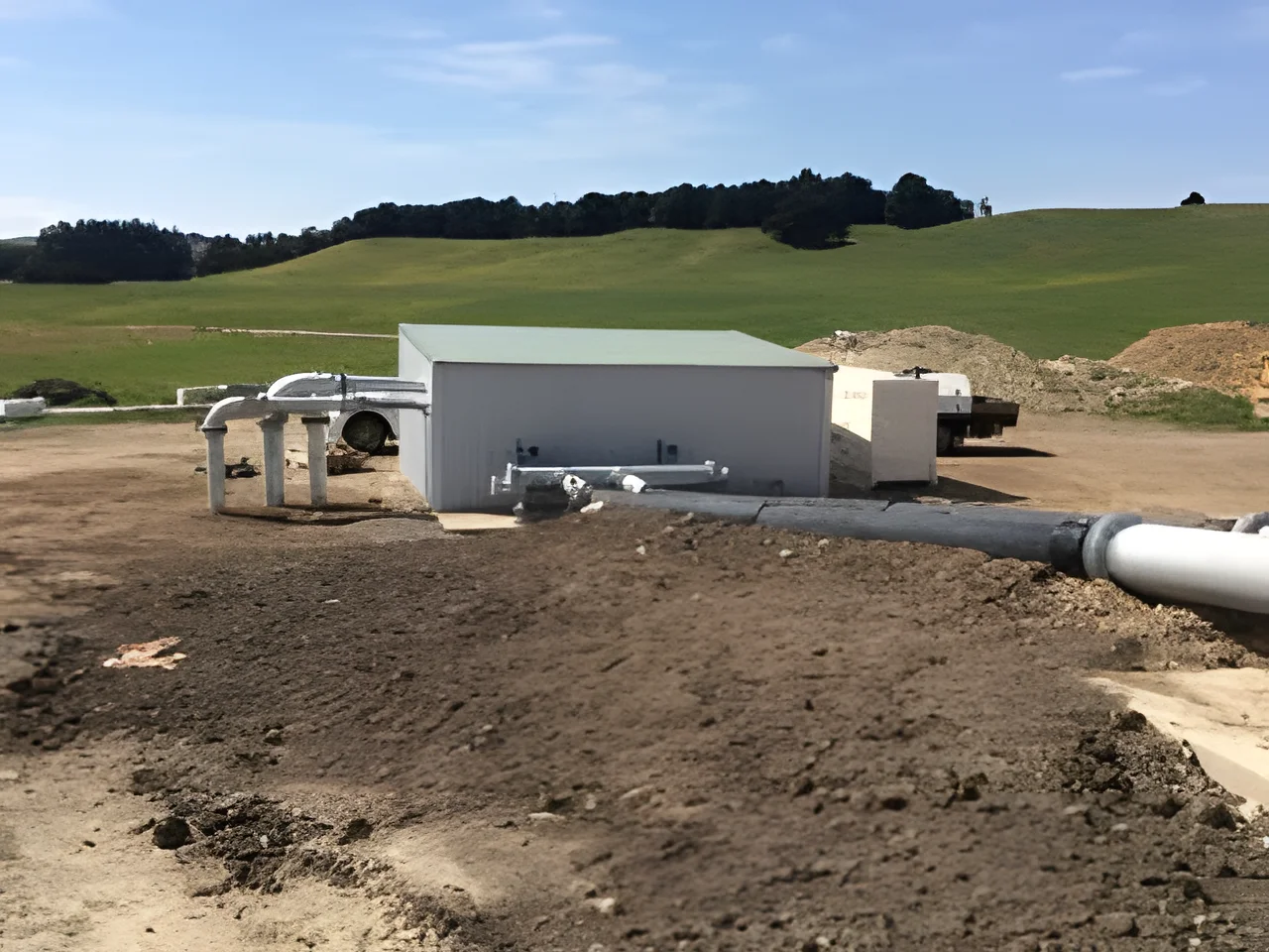 Large white pipework on stands running into a green-roofed shed on a farm, with a black pipe laid across freshly worked ground and green paddocks behind