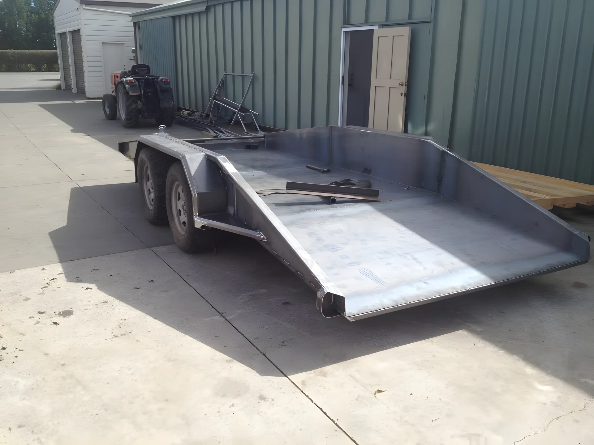 Steel transport trailer with a sloped deck, built and parked outside a workshop